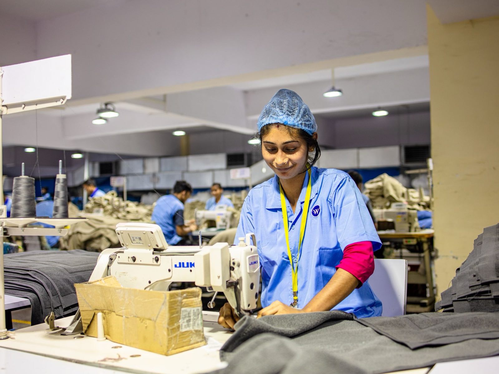 Machine operator in uniform and hair covering on a well-lit garment factory floor